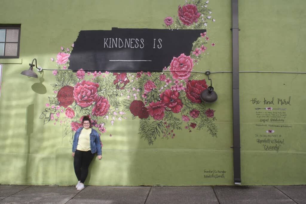 Dark digital wall sign with flowers, featuring "Kindness is..." message, mural art, and a smiling woman in front.