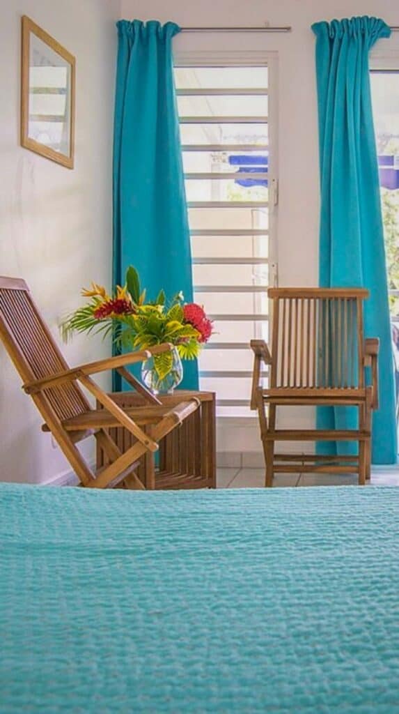 Bright les Cayalines guest room with teal curtains, wooden chairs, and a tropical flower arrangement
