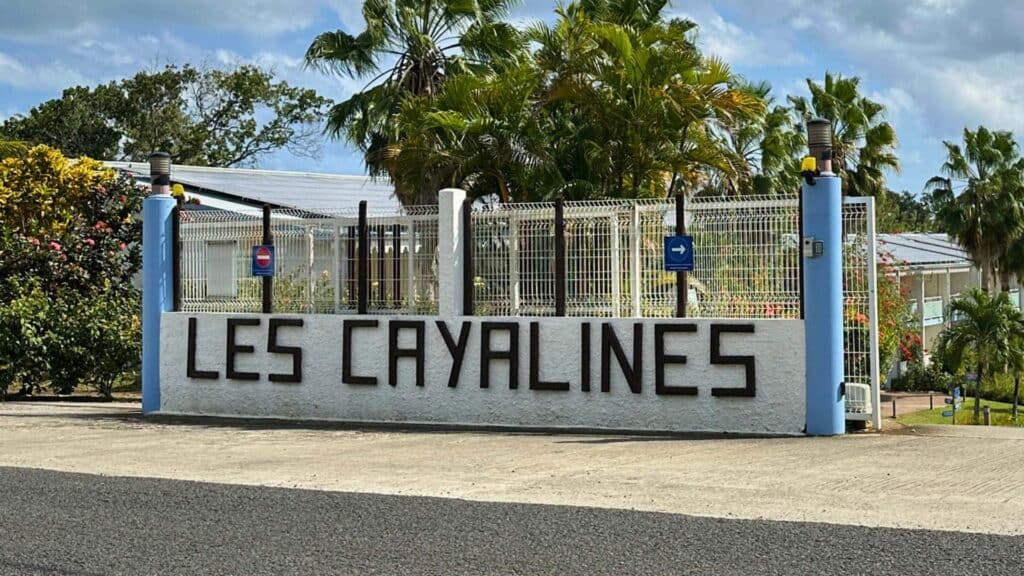 The Les Cayalines entrance sign with blue pillars, tropical trees, and flowering plants on a sunny day