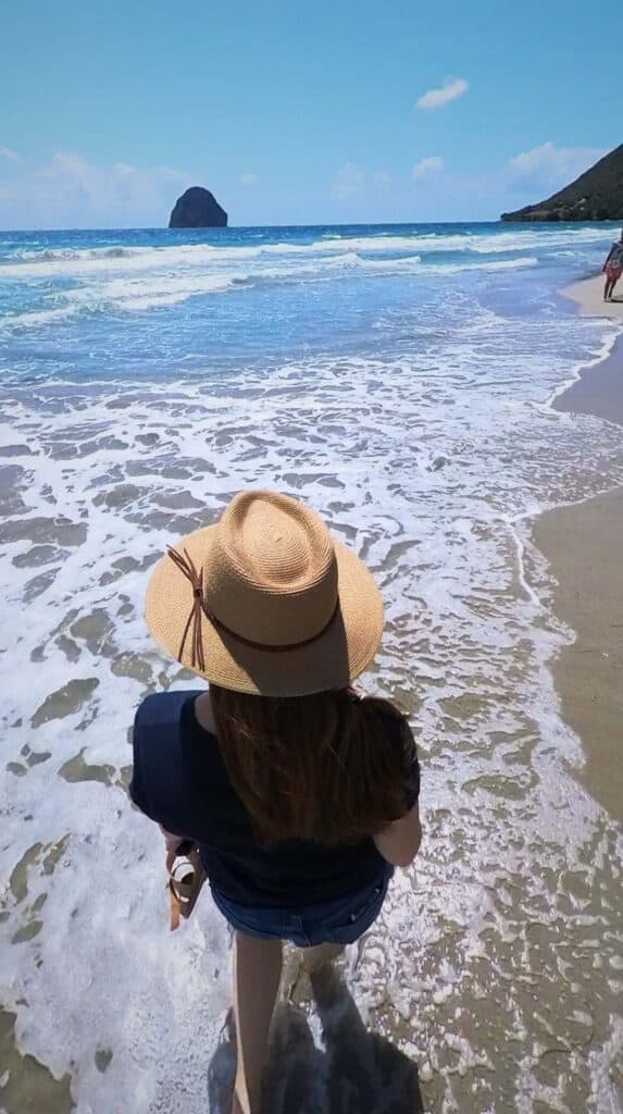 Woman in a sun hat standing at the water's edge on a Martinique beach with Diamond Rock in the distance