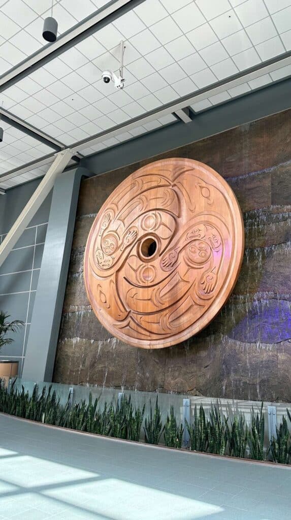 Large circular Indigenous art sculpture mounted on a wall inside Vancouver International Airport with a water feature below.