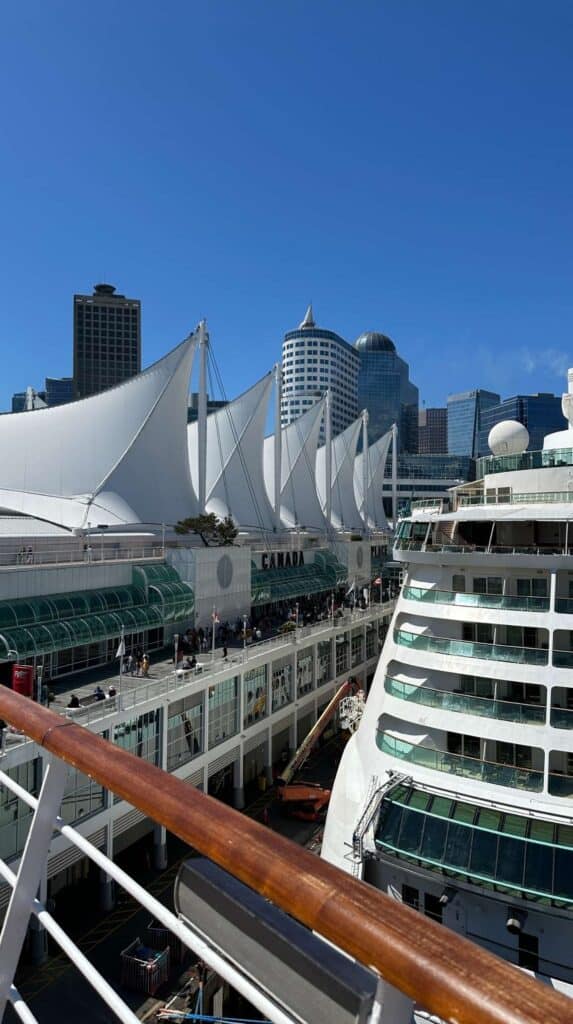 Cruise ship docked at Canada Place with white sail roof and downtown Vancouver skyline under a clear blue sky.