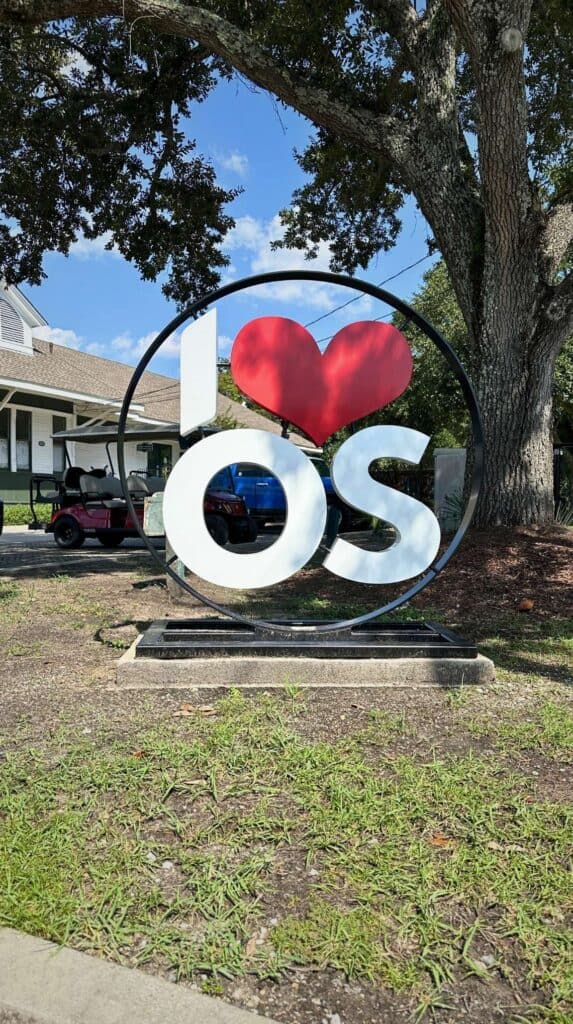 Large public art sign featuring a red heart and the letters "OS" displayed on a grassy area in Ocean Springs Mississippi near a large oak tree.