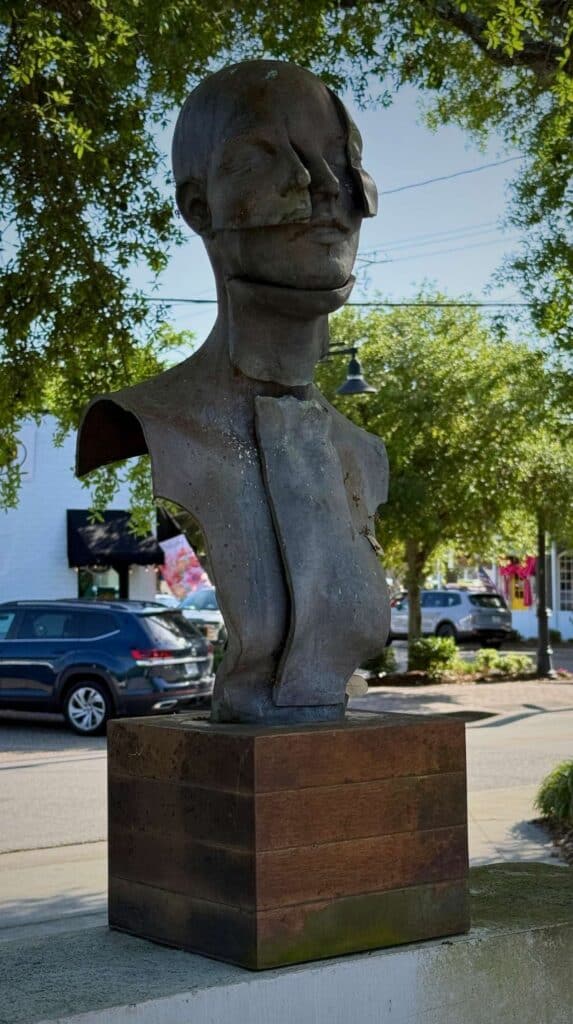 Large abstract bust sculpture displayed on a pedestal along a sidewalk in downtown Ocean Springs surrounded by trees and parked cars.
