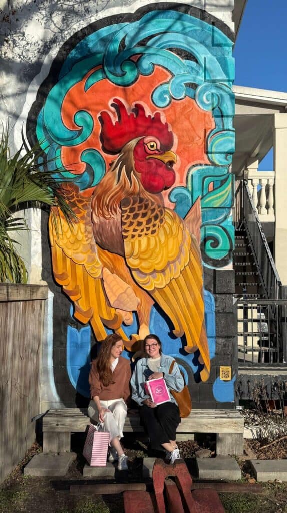 Large colorful mural of a rooster painted on a building wall with two people sitting in front of it in downtown Ocean Springs.