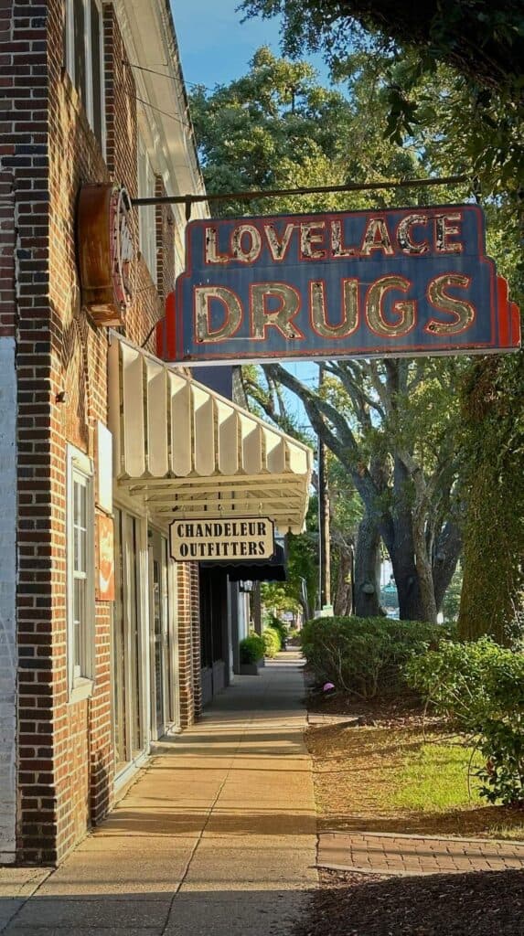 Colorful vintage sign reading "LOVELACE DRUGS" hanging above a shaded sidewalk in downtown Ocean Springs Mississippi with small local shops lining the street.