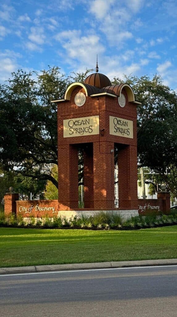 Brick welcome monument with arches and domed top displaying "Ocean Springs" beside a grassy roadside, marking the entrance to Ocean Springs Mississippi.