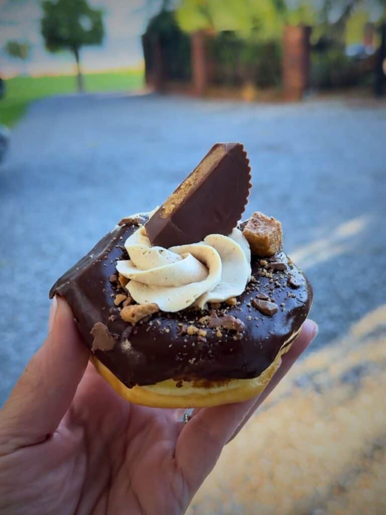 Hand holding a chocolate iced donut topped with peanut butter cup and cream, representing specialty sweets at Ocean Springs restaurants.