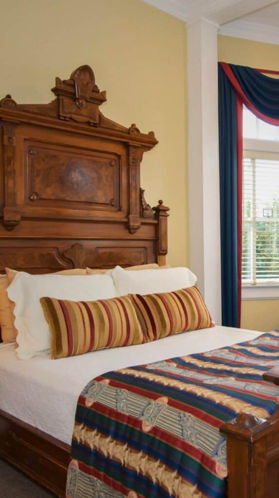 Historic guest bedroom at Bluff View Inn with carved wooden headboard, striped bedding, and tall windows with draped curtains.
