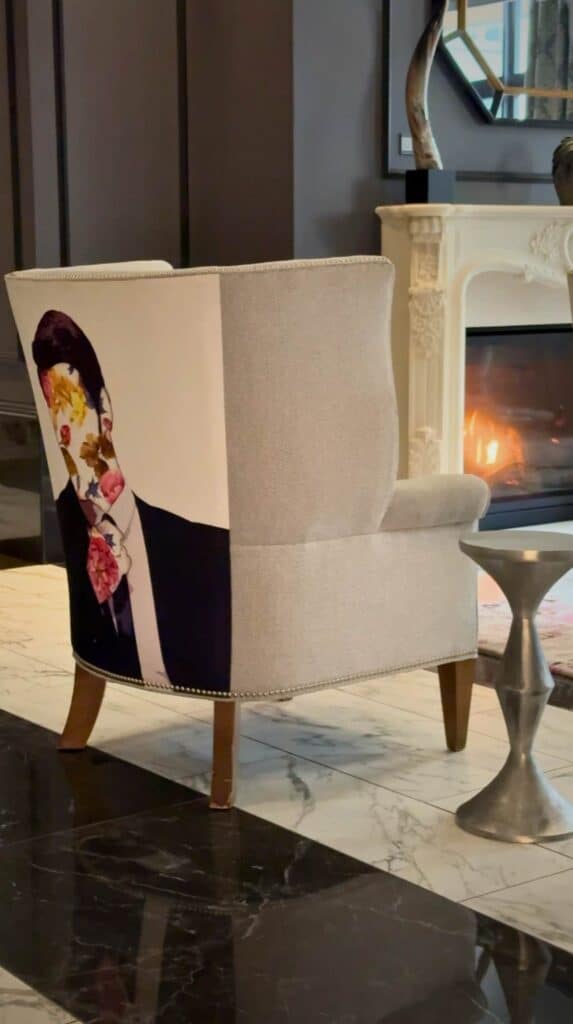 White accent chair with floral artwork beside a fireplace inside The Edwin Hotel showcasing the artistic design style of this unique place to stay in Chattanooga.