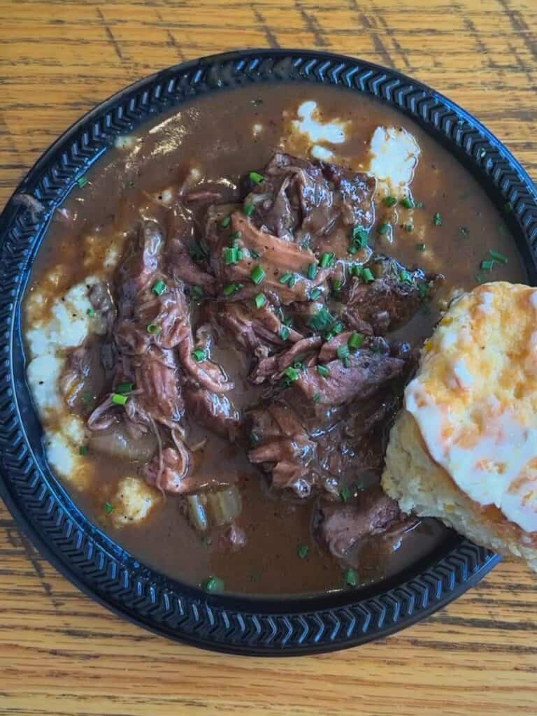 Braised beef shank covered in rich brown gravy and topped with chopped herbs on a black plate. The dish is served over mashed potatoes with a biscuit on the side at one of the best restaurants in Ocean Springs.