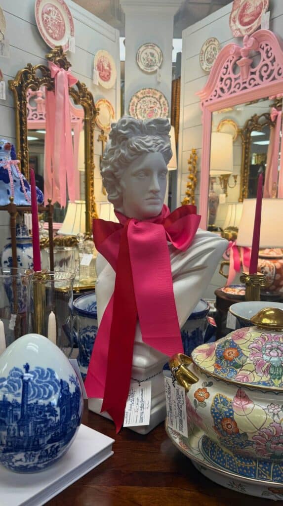 Vintage decor display inside Cottage Grove Vintage in Greenville featuring a white bust statue tied with a bright pink ribbon surrounded by patterned ceramics and mirrors.