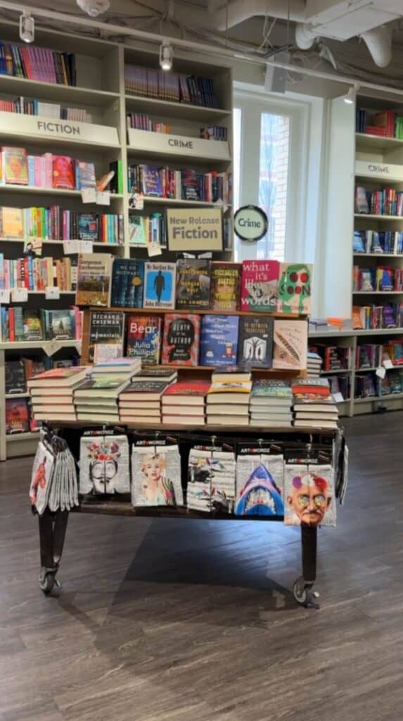 Central display table covered with books and gifts inside M. Judson Booksellers surrounded by shelves of fiction and art books.