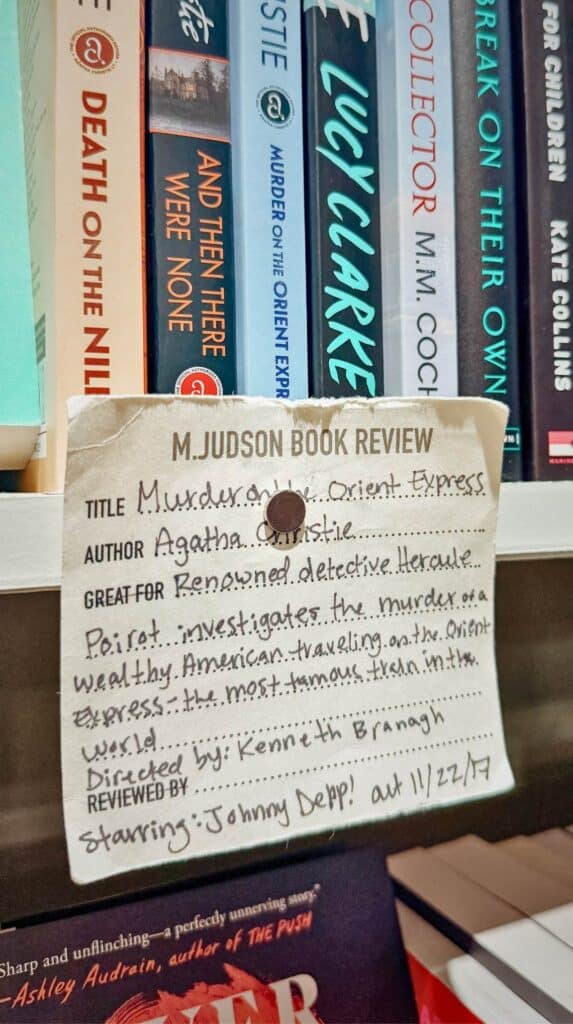 Handwritten “M. Judson Book Review” recommendation card displayed in front of mystery novels on a bookstore shelf at M. Judson Booksellers.