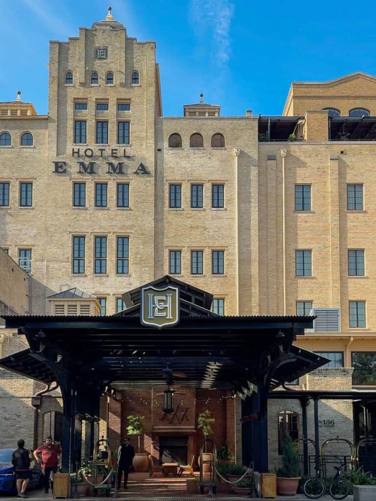 The stone facade and covered entrance of Hotel Emma San Antonio against a bright blue sky
