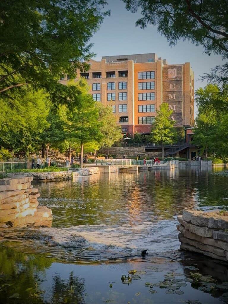 The brick Emma tower of Hotel Emma San Antonio reflected in the river and framed by green trees