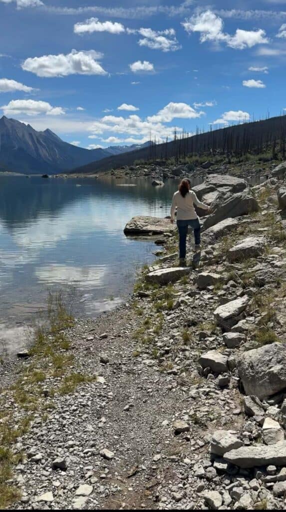 Person walking along a rocky shoreline beside calm water with mountains in the distance near Maligne Lake.