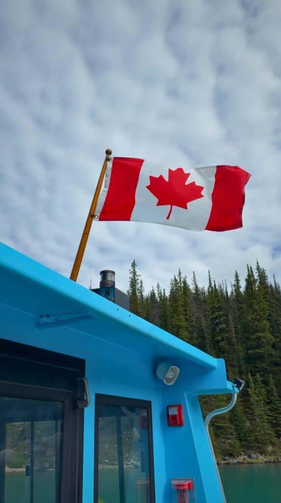 Canadian flag flying above a blue boat with evergreen trees and cloudy sky at Maligne Lake.