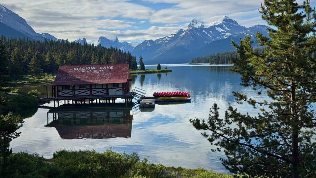 "Maligne Lake Boat House" building on calm water with canoes docked nearby and snow capped mountains reflected in the lake.