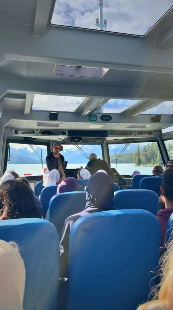 Passengers seated inside a glass roof boat during a Maligne Lake cruise while a guide speaks at the front with lake and mountains visible ahead.