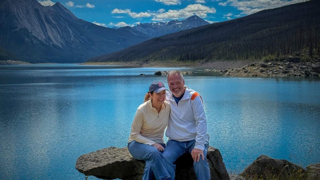 Couple sitting on a rock with turquoise Medicine Lake and forested mountains behind them on a clear day.