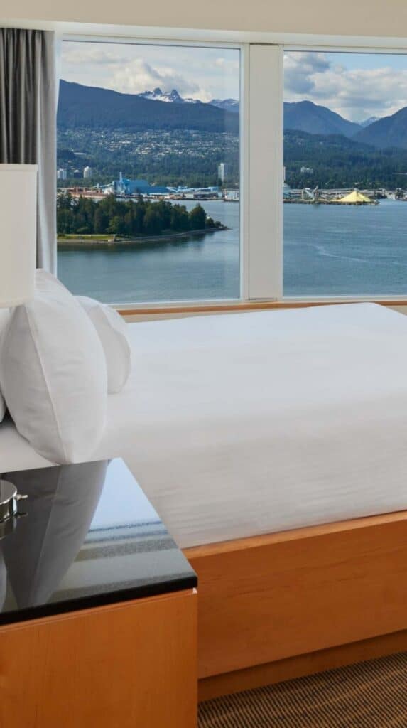 Hotel room with bed facing large windows overlooking Vancouver harbor and mountains near the cruise port.