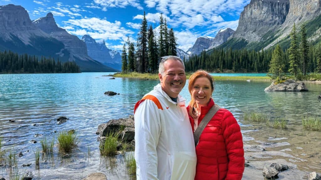 Couple smiling at the camera in front of Spirit Island Maligne Lake with turquoise water pine trees and dramatic mountain peaks behind them.