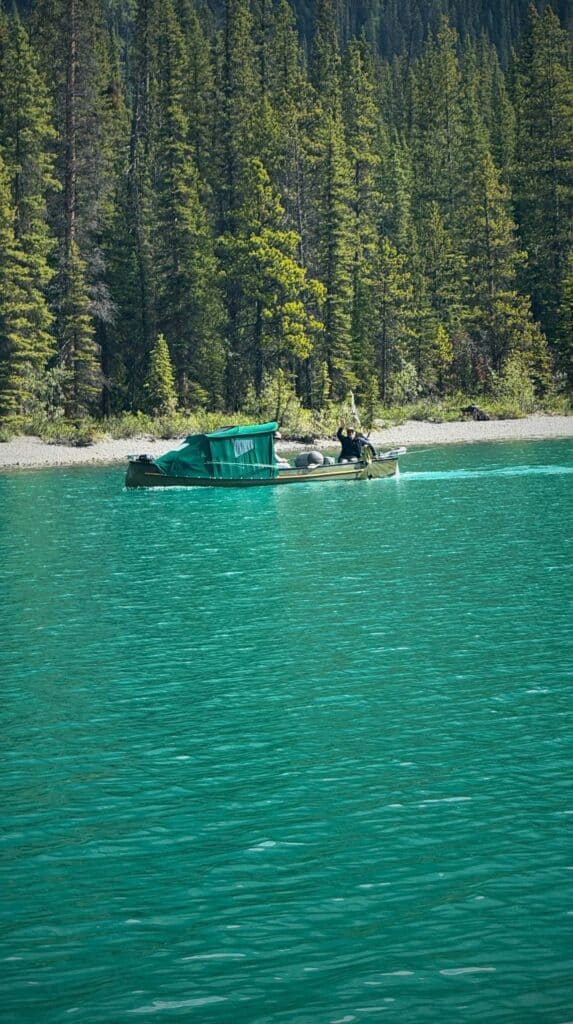 Small boat cruising across turquoise Maligne Lake with a person onboard raising an arm near a forested shoreline.