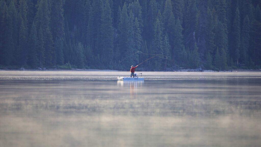 Fly fisherman standing in calm water with forest and mountains behind him on Maligne Lake.