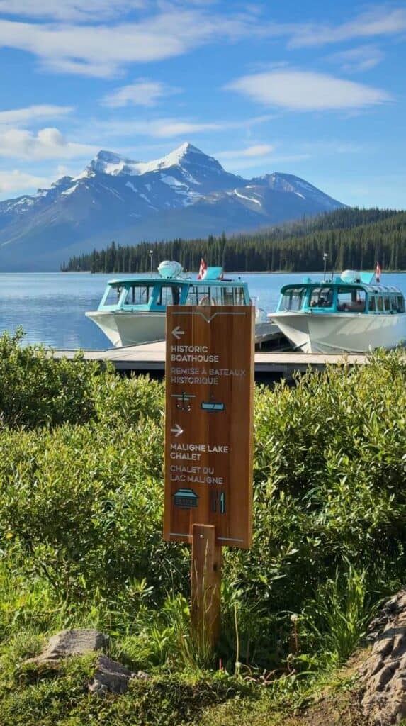 Wooden sign pointing to "Historic Boathouse" and "Maligne Lake Chalet" with boats and mountains behind it.