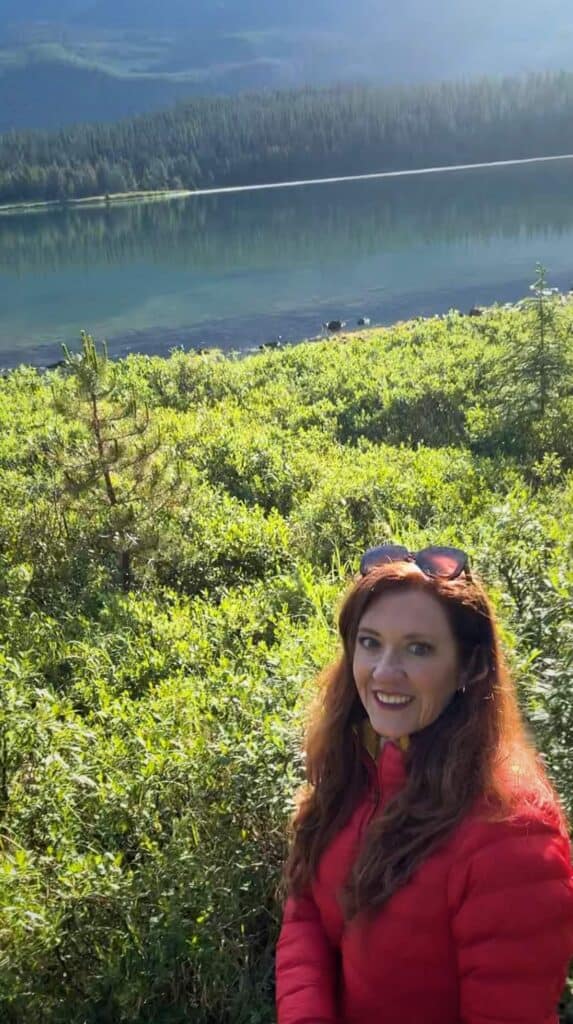Woman in a red jacket smiling near the grassy shoreline of Maligne Lake with calm water and forested hills in the background.