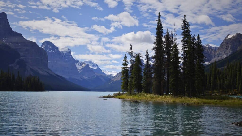 Spirit Island Maligne Lake with tall pine trees on a small island surrounded by calm water and dramatic mountain peaks under a partly cloudy sky.