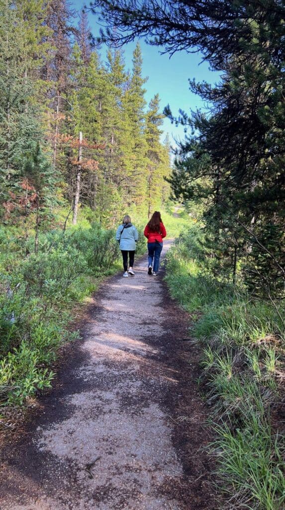 Two people walking along a forest trail surrounded by tall pine trees near Maligne Lake on a sunny day.