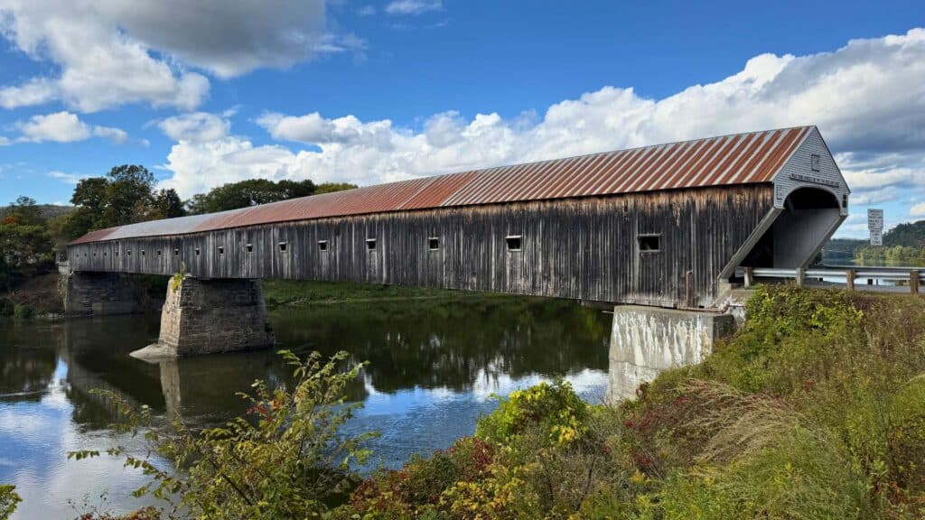 A weathered covered bridge reflects in the still river near Woodstock, Vermont