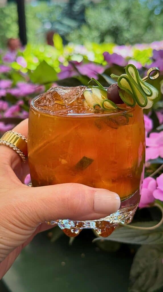 A cucumber-garnished cocktail held against pink patio flowers at the Broadmoor Golden Bee