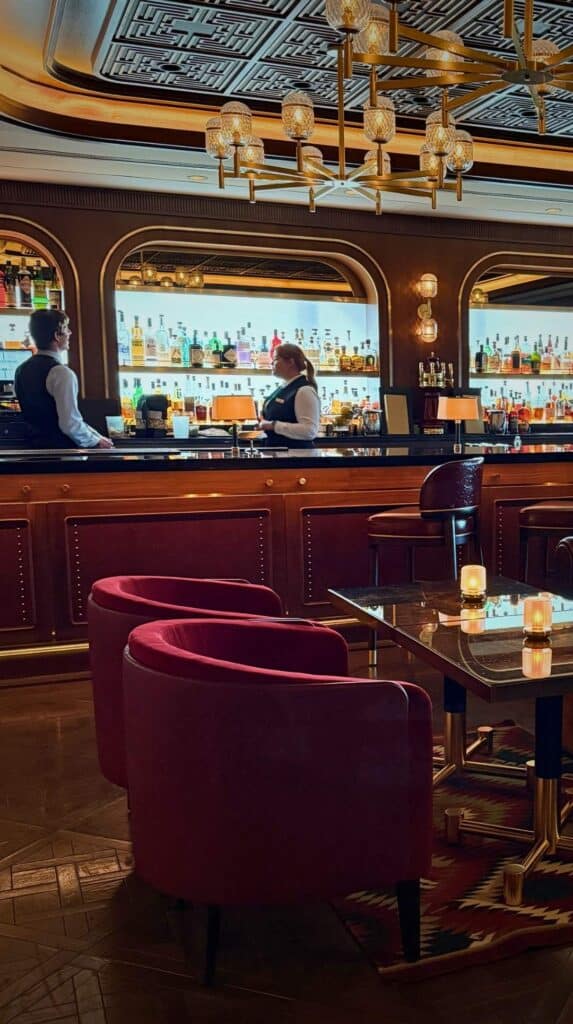 Rich red velvet chairs and a glowing back bar under a gilded ceiling at the Penrose Lounge