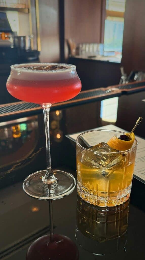 A pink coupe cocktail and an orange-twist old fashioned on the black bar at the Penrose Lounge